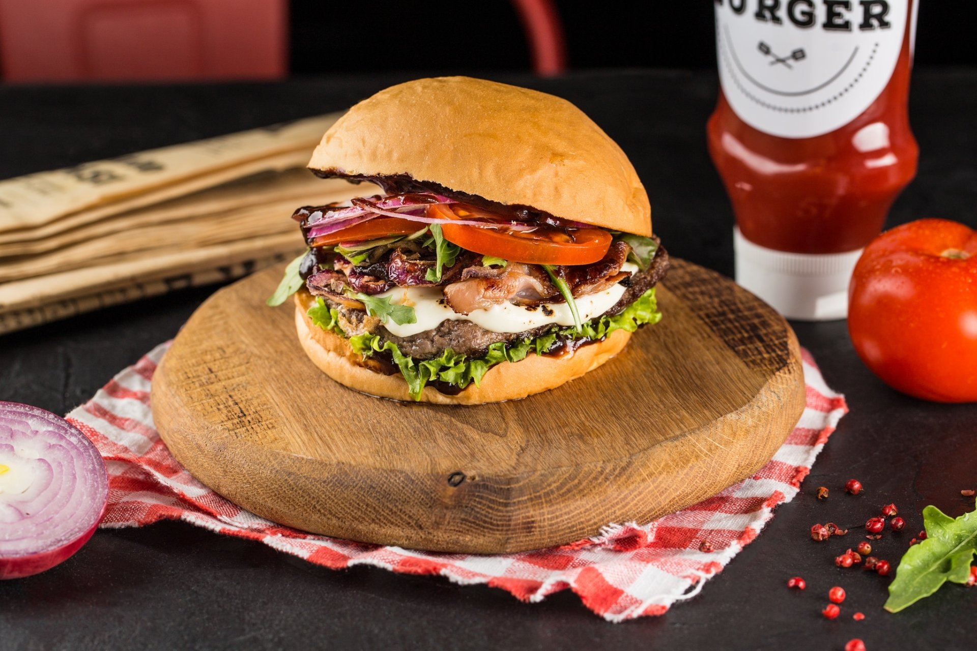 HD PC desktop wallpaper still life of a juicy burger on a wooden board with lettuce, tomato, red onion and a ketchup bottle, vibrant food background.