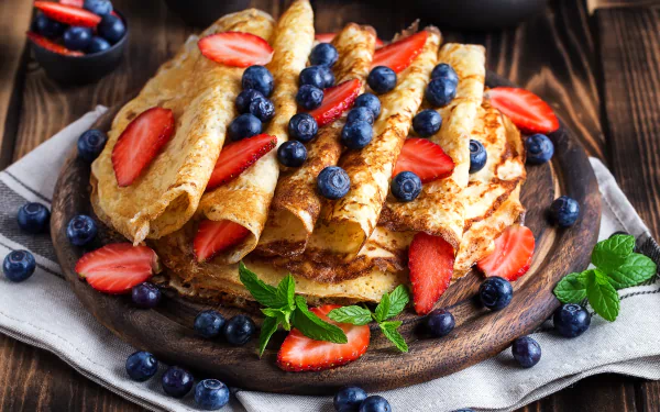 A 4K Ultra HD still life of crêpes topped with fresh strawberries and blueberries, garnished with mint leaves on a rustic wooden table.