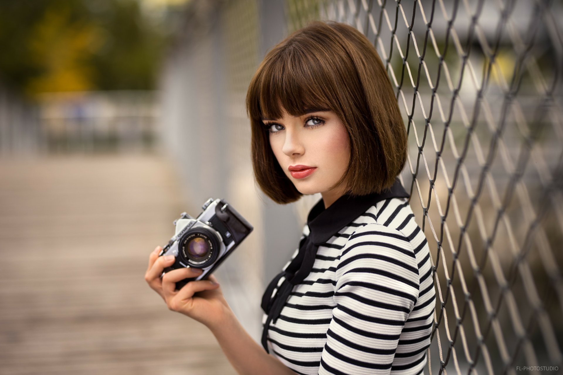 HD desktop wallpaper of a brunette model with short hair and striking blue eyes holding a vintage camera beside a chain-link fence.