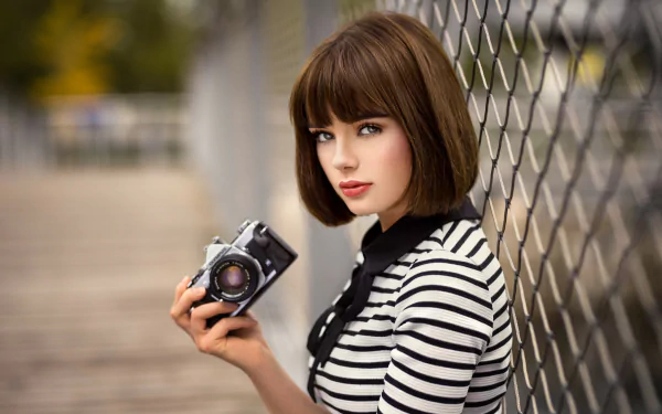HD desktop wallpaper of a brunette model with short hair and striking blue eyes holding a vintage camera beside a chain-link fence.