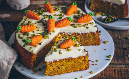 HD desktop wallpaper featuring a moist carrot cake topped with cream cheese frosting, miniature carrot decorations, and chopped pistachios on a white plate.