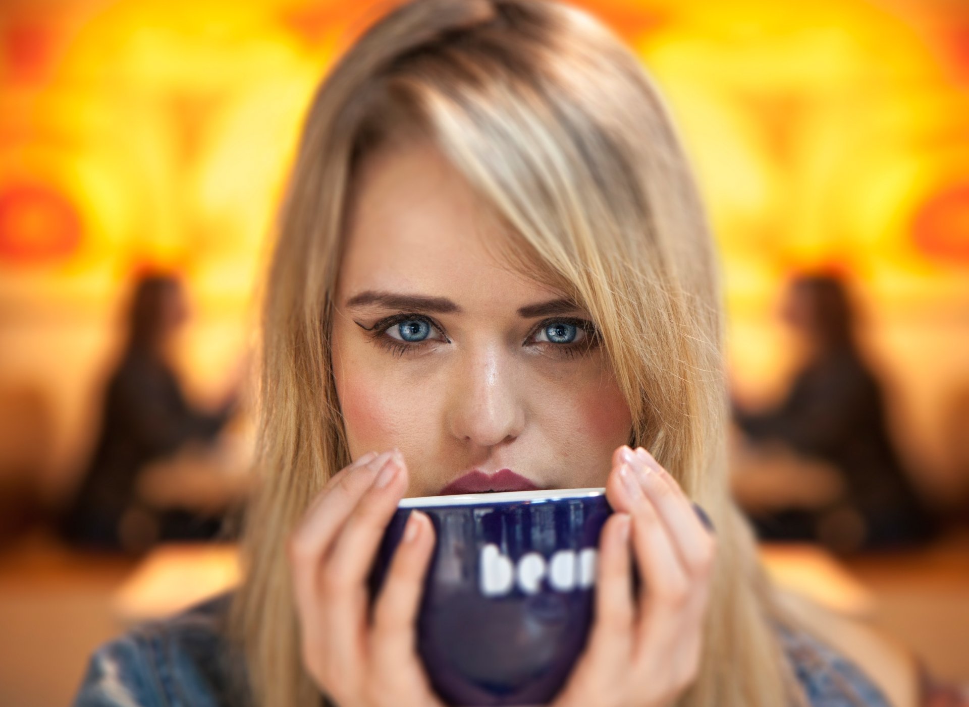 4K PC desktop wallpaper: close-up face of a blonde woman model with blue eyes, holding a mug to her mouth, intense gaze and soft warm bokeh background.