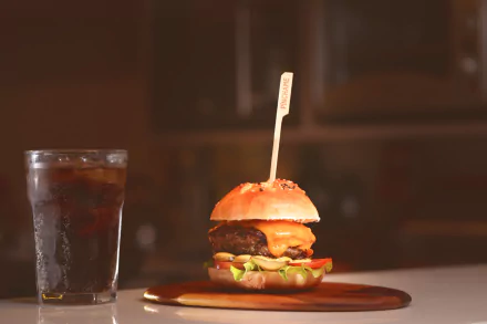 4K Ultra HD wallpaper featuring a juicy hamburger with melted cheese and fresh lettuce, accompanied by a cold drink on a wooden surface against a blurred background.