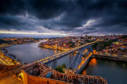 Night view of the Luís I Bridge spanning the river in Porto, Portugal, with city lights glowing under a dramatic cloudy sky.