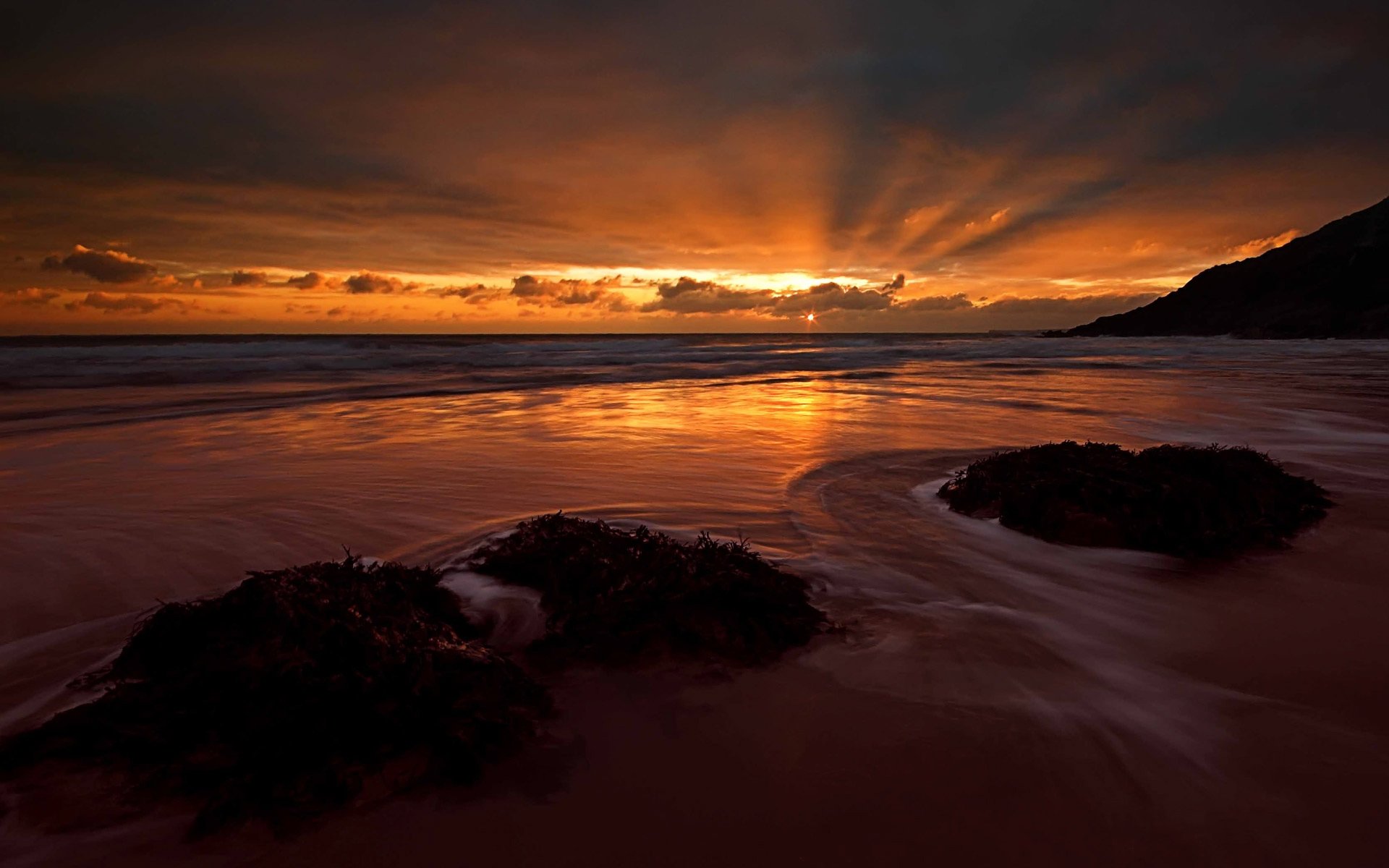 HD PC desktop wallpaper of a dramatic sunset over the ocean horizon, sunbeams through clouds reflecting on wet sand, seaweed on the beach and a silhouetted headland.