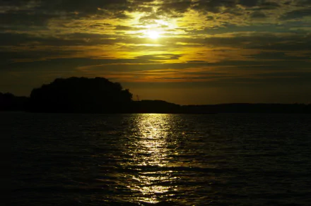 HD desktop wallpaper of a serene lake at sunset, with golden sunlight reflecting on the water under a partly cloudy sky surrounded by nature.