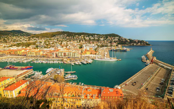 A vibrant 4K Ultra HD view of Nice, France, showcasing a bustling marina with boats, a sprawling cityscape, and the calm Mediterranean coastline under a partly cloudy sky.