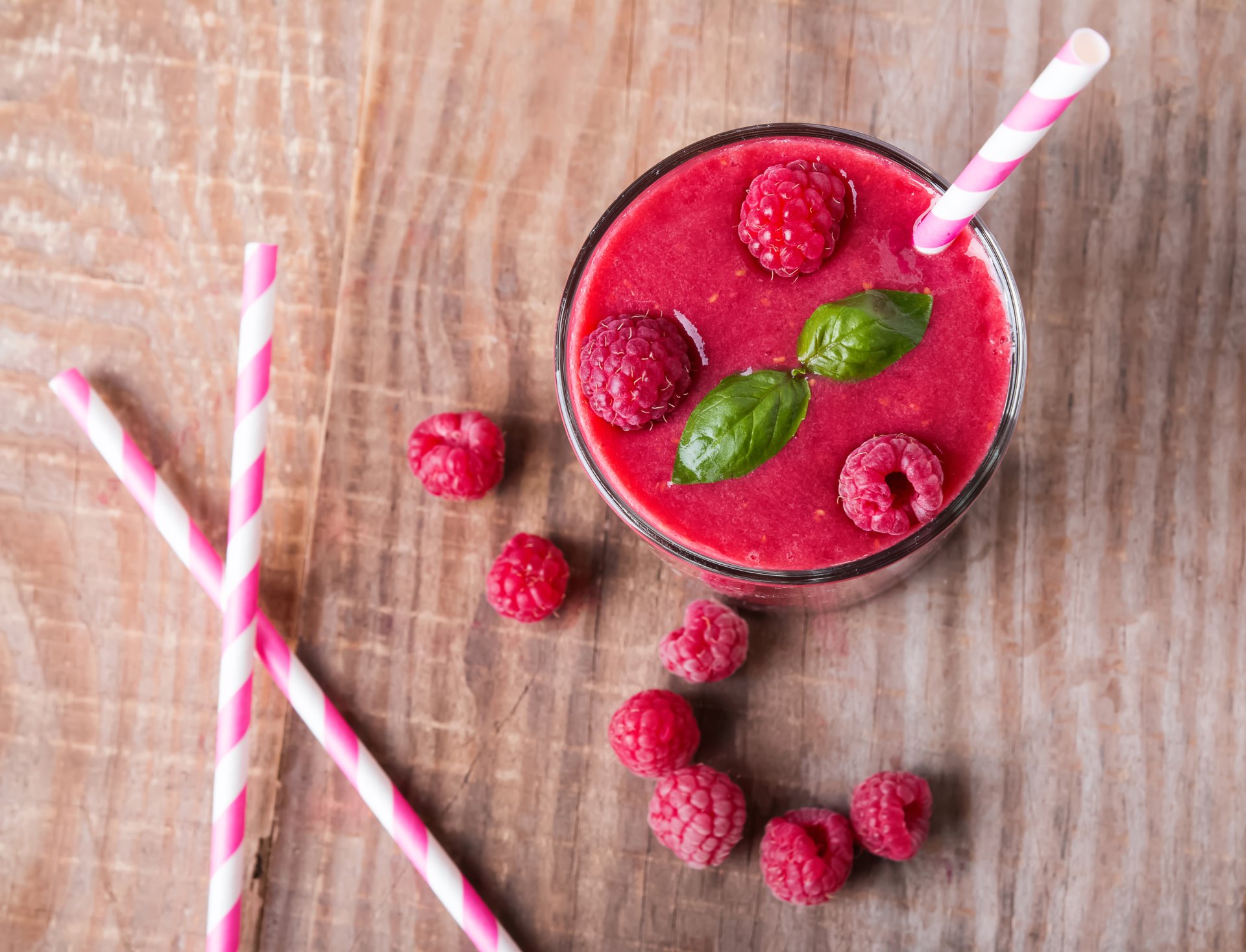 4K Ultra HD still-life of a raspberry smoothie in a glass with basil leaves and a striped straw, scattered raspberries on a wooden background — food/drink PC desktop wallpaper.