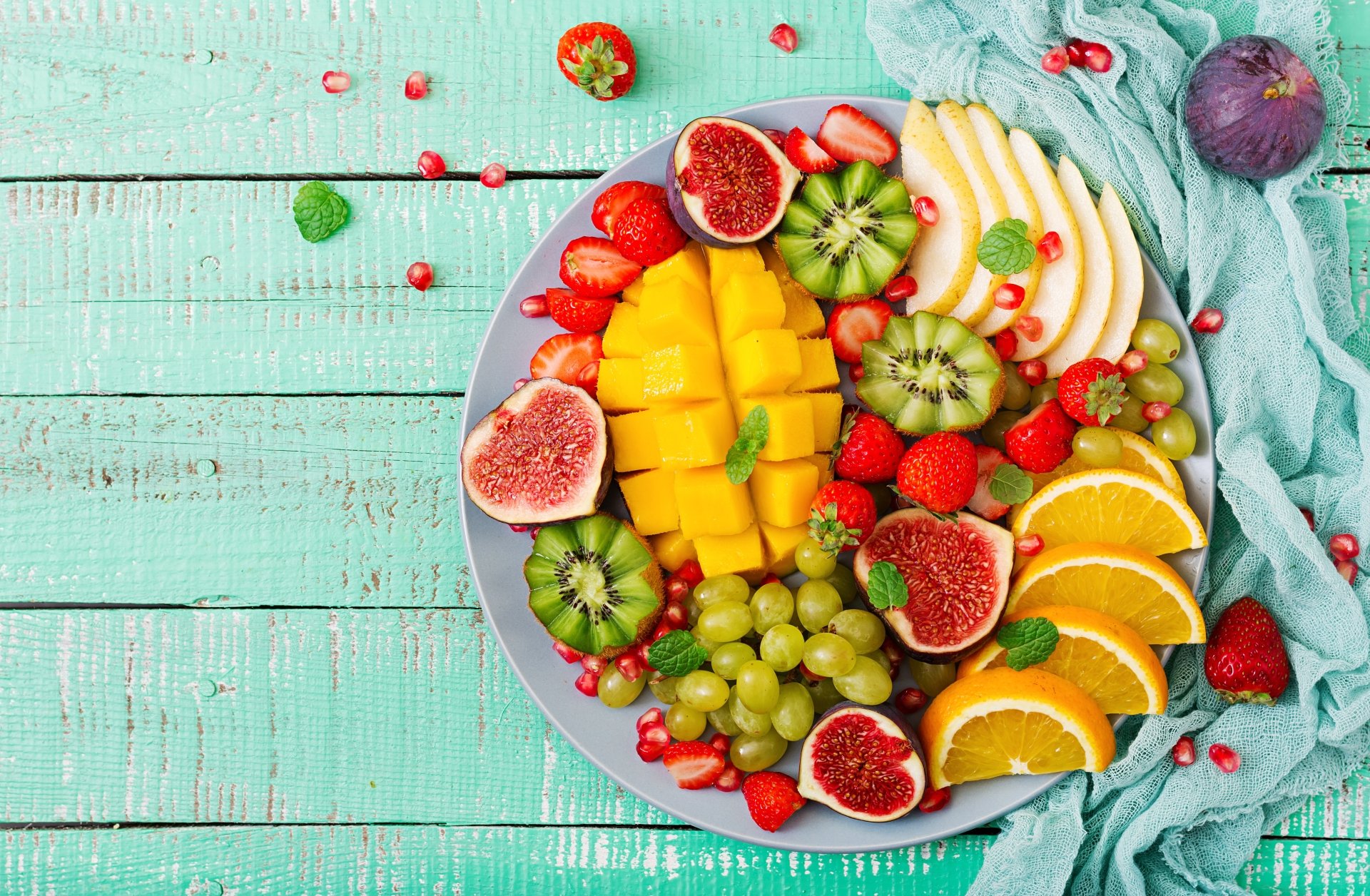 Vibrant still life of fresh strawberries, grapes, figs, kiwi, mango, and citrus on a blue plate, displayed on a turquoise wooden surface in 4K Ultra HD.