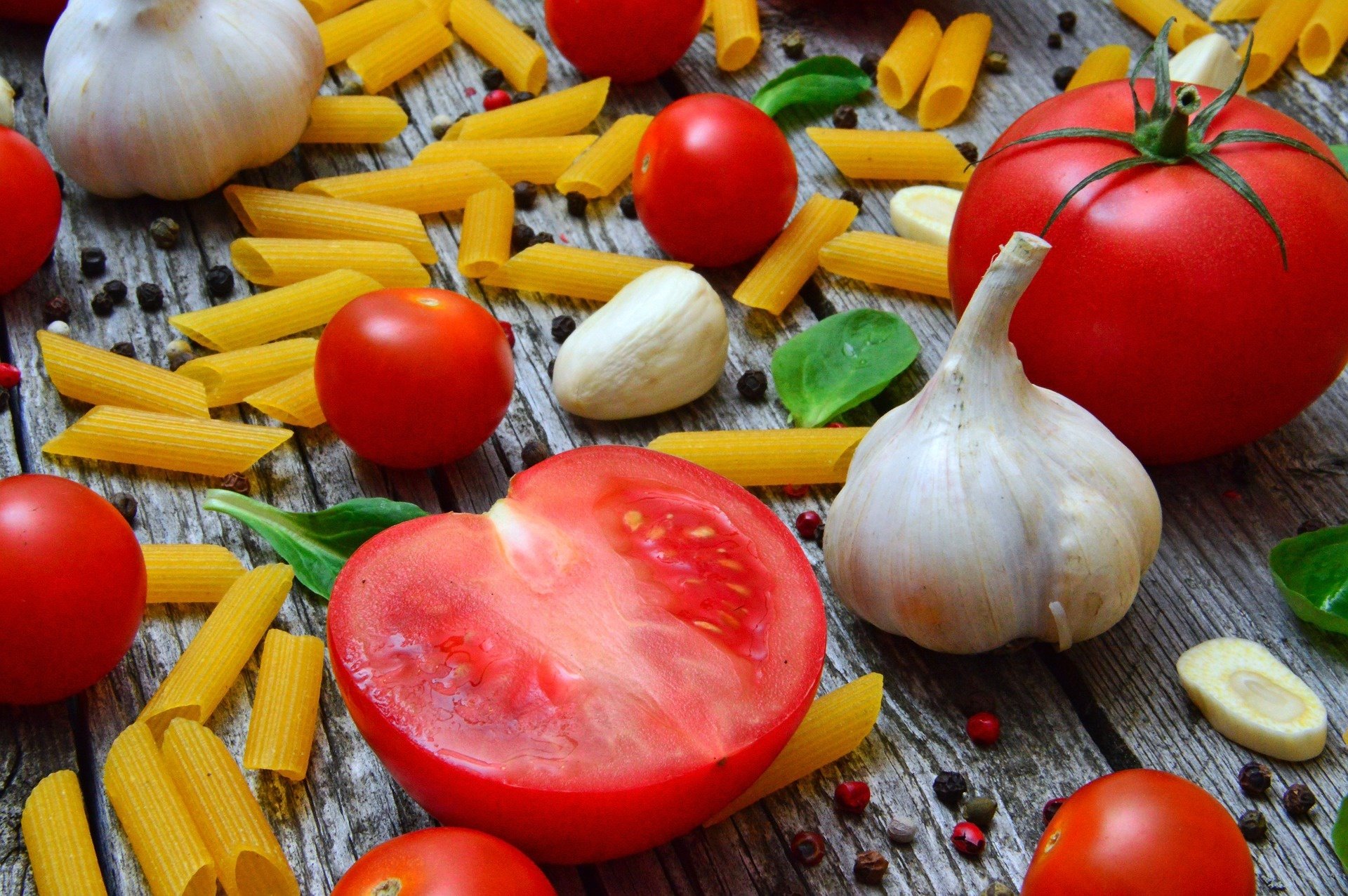 HD PC desktop wallpaper featuring a still life of garlic cloves, fresh tomatoes, cherry tomatoes, and uncooked pasta scattered on a rustic wooden surface.