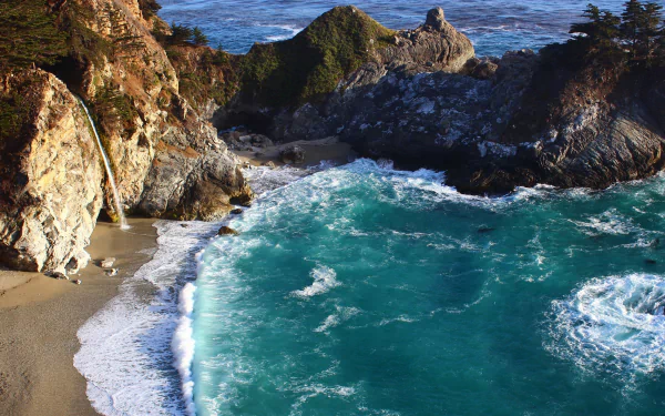HD desktop wallpaper of McWay Falls in Big Sur, California, showing the waterfall cascading down a cliff onto a pristine beach by the vibrant, turquoise coastal waters. Lush greenery tops the rugged cliffs.
