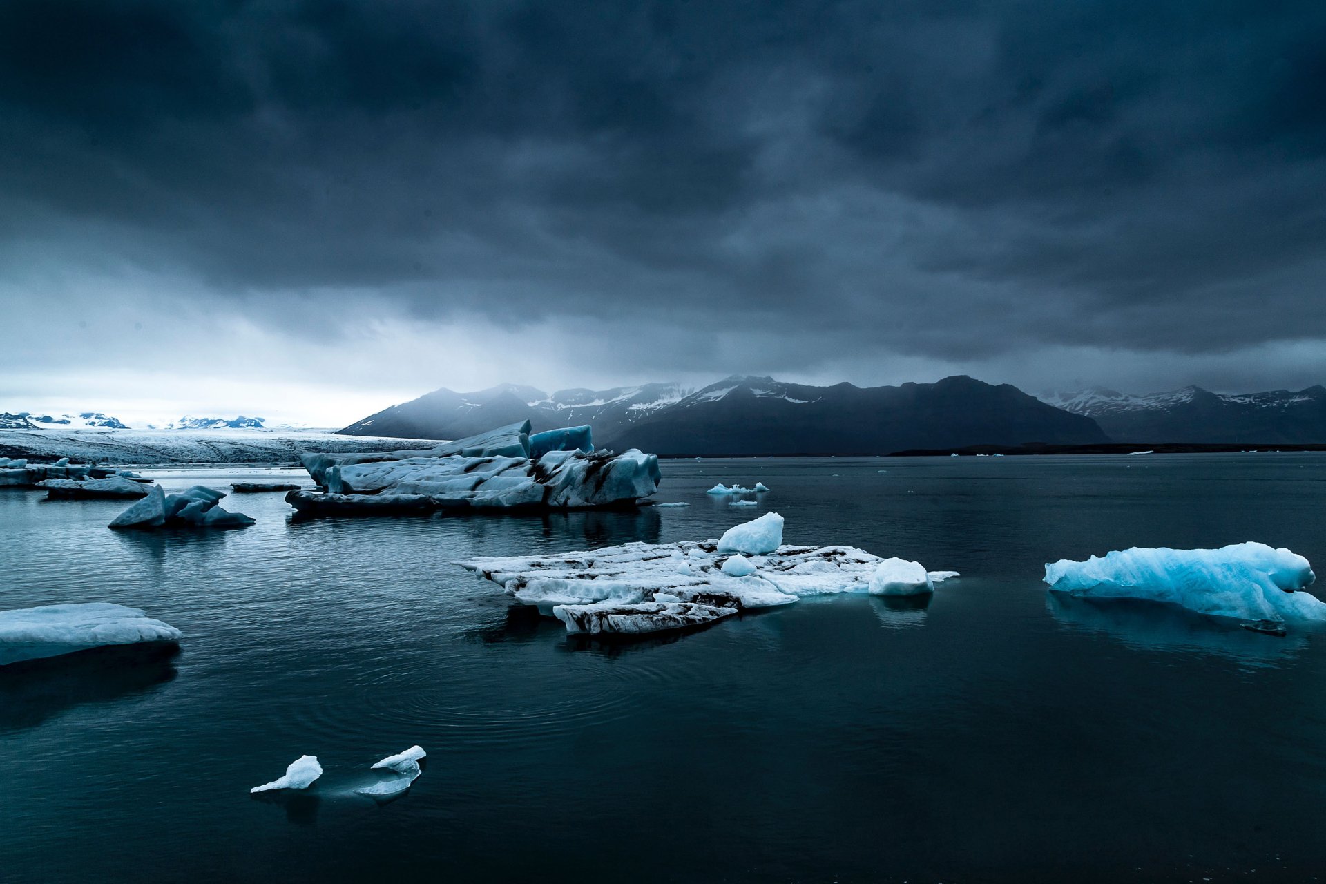 HD wallpaper featuring a serene nature scene with icebergs floating in calm dark blue water, set against a backdrop of mountains under an overcast sky. An ideal background for desktops.