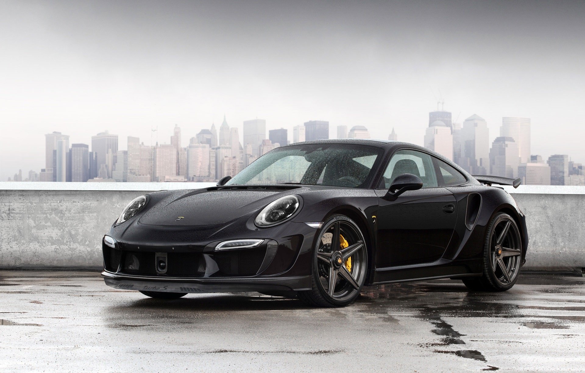 Black Porsche 911 sports car parked on a wet rooftop with a hazy city skyline, HD PC desktop wallpaper background.