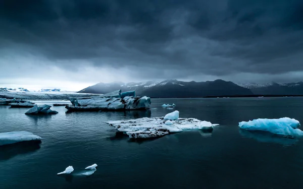 HD wallpaper featuring a serene nature scene with icebergs floating in calm dark blue water, set against a backdrop of mountains under an overcast sky. An ideal background for desktops.