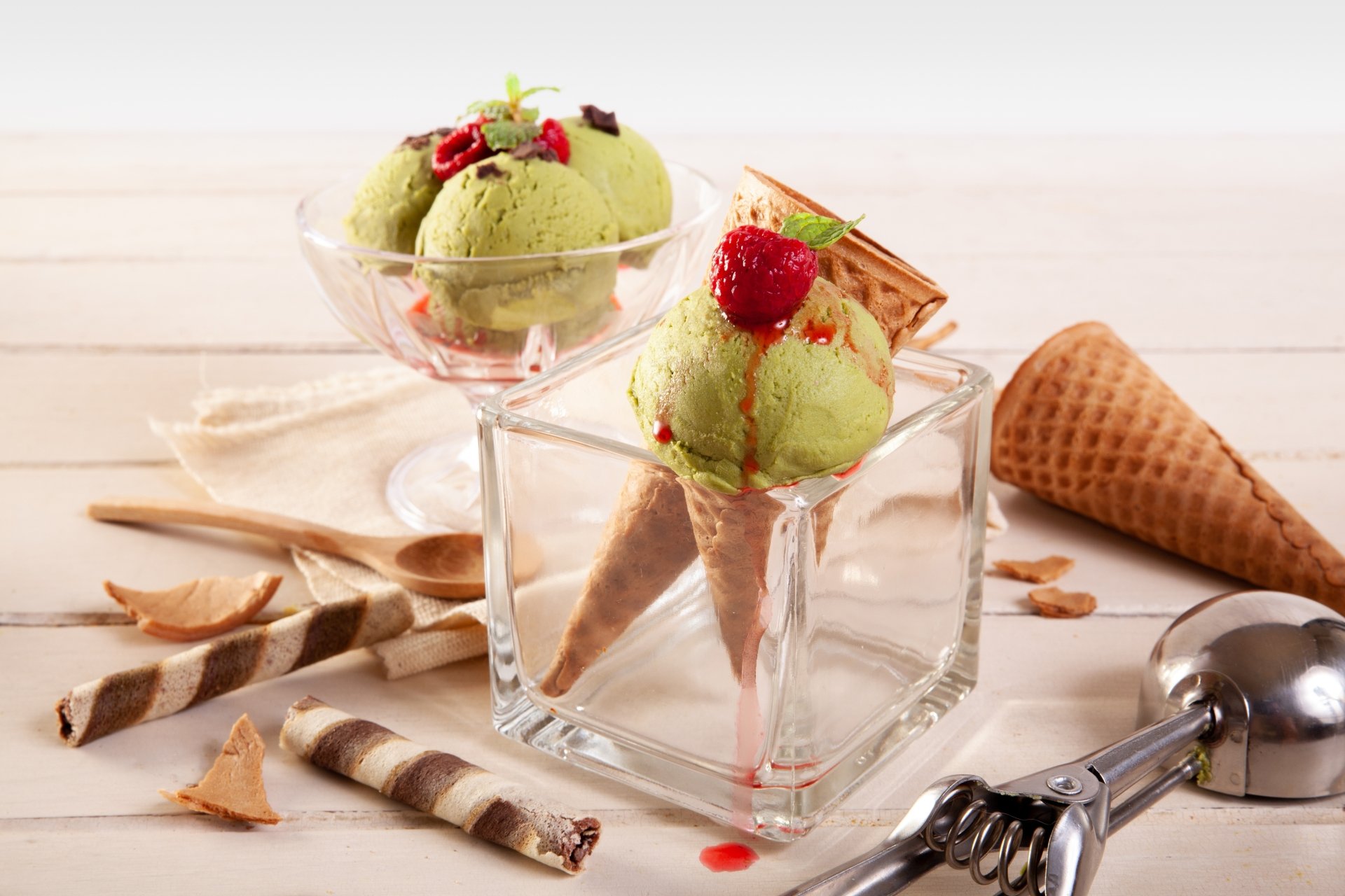Still life of pistachio-green ice cream scoops in waffle cones set in glass dishes with wafer rolls and a spare cone on a wooden table — 5K Ultra HD PC desktop wallpaper.