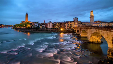 Evening view of the river and historic man-made bridge in Verona, Italy, with illuminated city buildings under a moody sky.