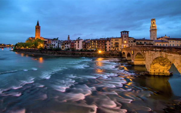 Evening view of the river and historic man-made bridge in Verona, Italy, with illuminated city buildings under a moody sky.