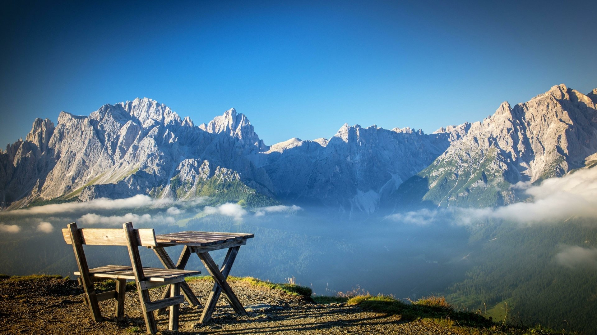 A wooden chair and table set on a mountaintop overlooking clouds and rugged peaks, captured in vibrant 4K Ultra HD photography for a desktop wallpaper background.