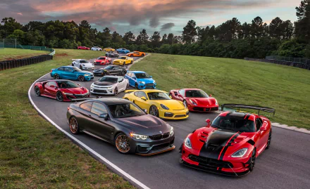 A lineup of high-performance supercars including Lexus, Jaguar, BMW, Audi, Acura, Ferrari, Dodge, Porsche, Ford, and McLaren on a racetrack at sunset.