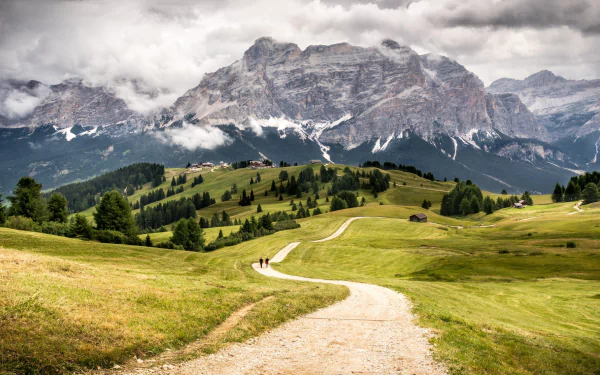 A winding path leads through grassy hills towards the towering Dolomites mountains in Italy under a cloudy sky, captured in stunning HD photography.