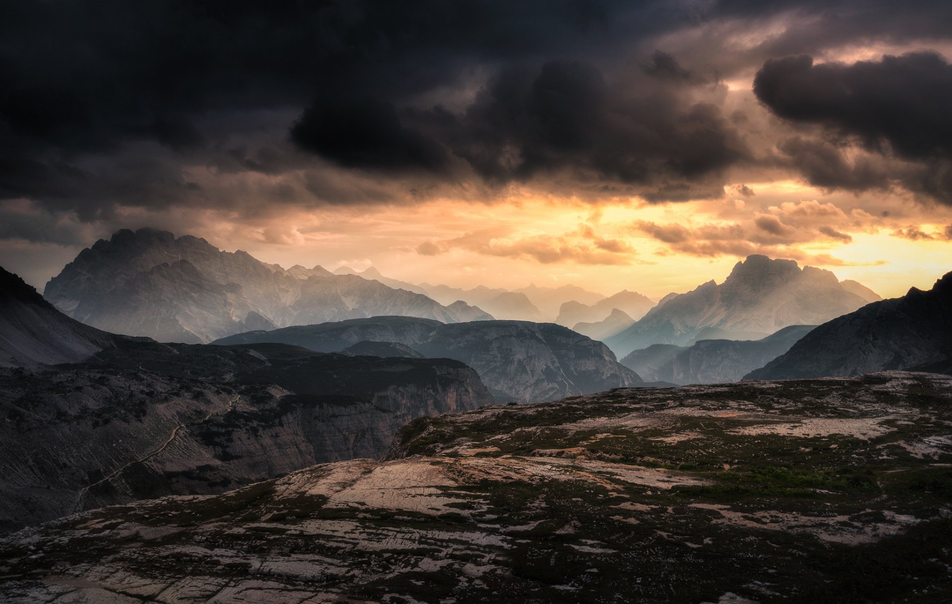 HD PC desktop wallpaper: dramatic mountain landscape and rugged foreground under heavy storm clouds, golden light spilling through distant peaks — a moody nature scene.