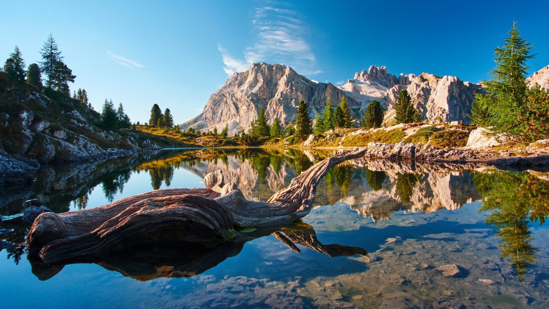 HD desktop wallpaper featuring a serene mountain landscape with a clear lake reflecting rugged peaks and surrounding trees under a bright blue sky.