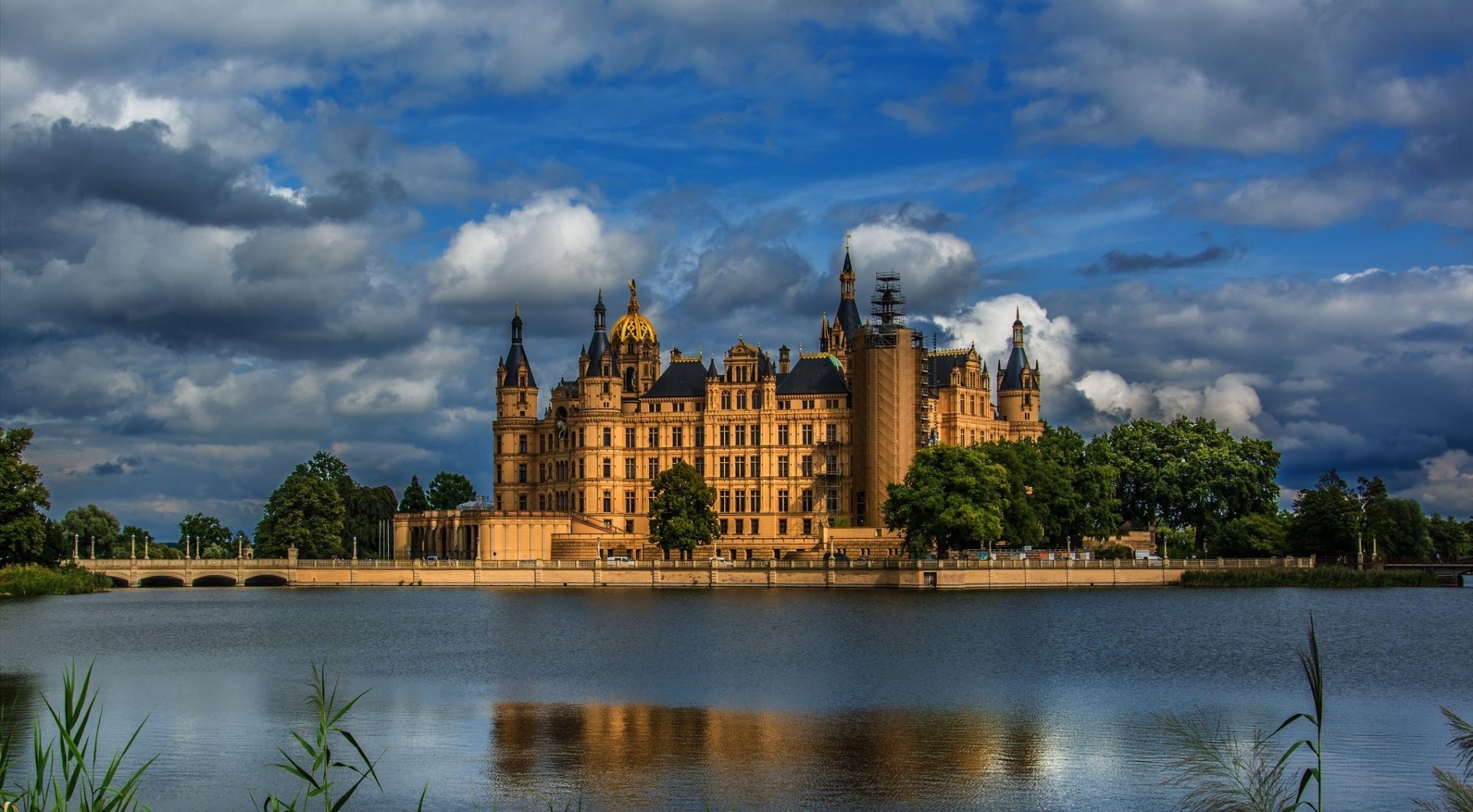HD desktop wallpaper featuring Schwerin Palace, a grand man-made historic castle set against a dramatic cloudy sky, reflected in the calm waters below.