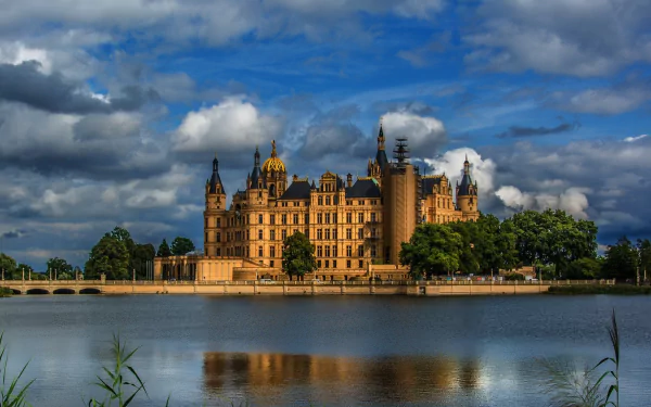 HD desktop wallpaper featuring Schwerin Palace, a grand man-made historic castle set against a dramatic cloudy sky, reflected in the calm waters below.