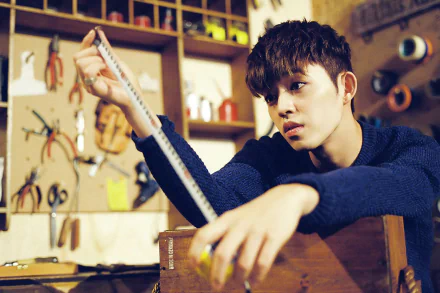 HD PC wallpaper of a young man in a workshop, holding a ruler and gazing pensively amid tools and warm lighting, with a subtle music-themed atmosphere.