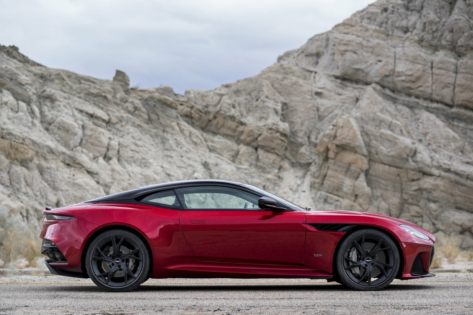 A red Aston Martin DBS Superleggera supercar parked against a rocky mountainous backdrop, featured as a HD PC desktop wallpaper and background.
