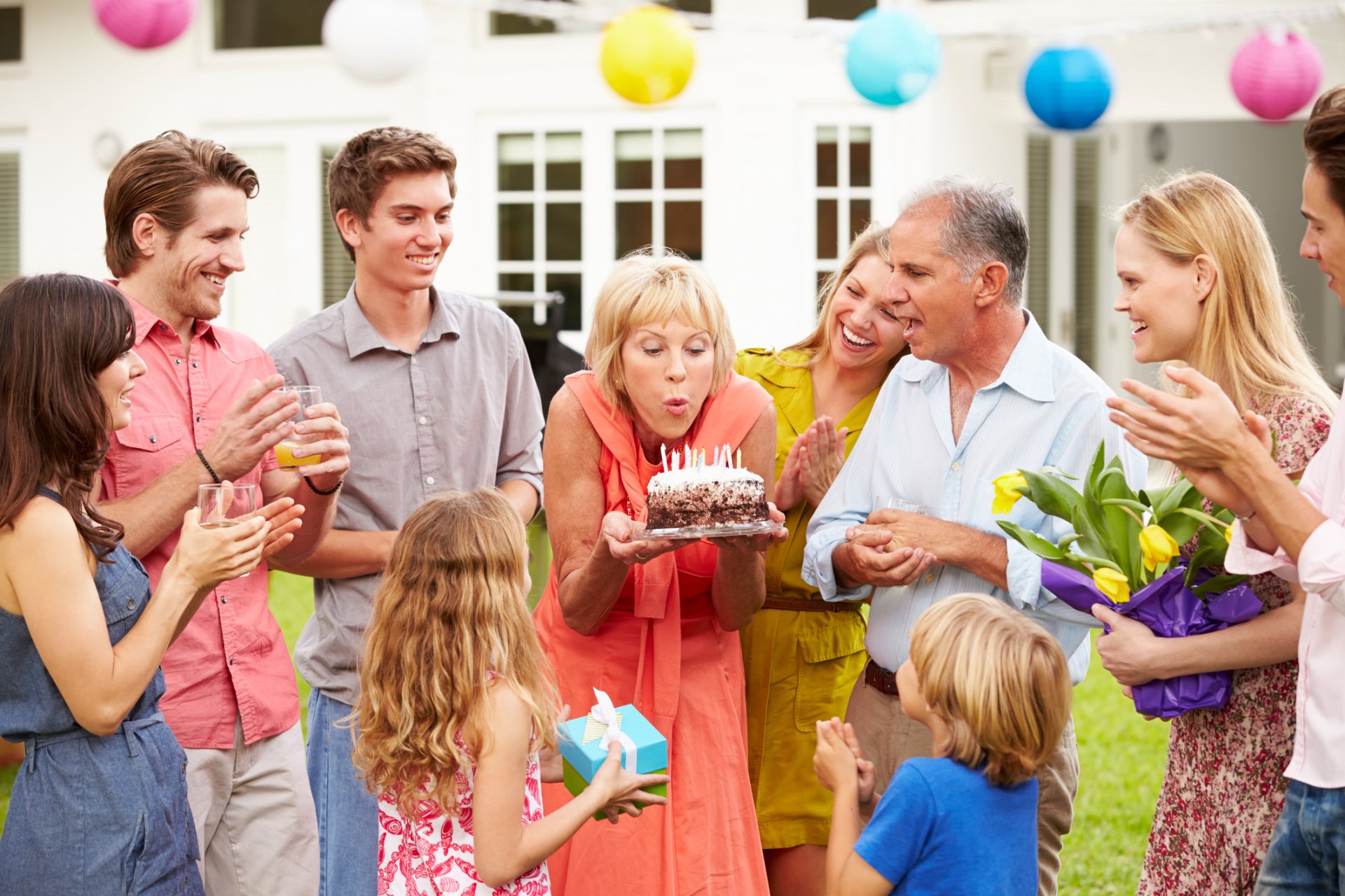 Holiday birthday celebration: multi-generation family in a backyard with balloons, woman blowing out cake candles as children and adults smile — HD PC desktop wallpaper/background.