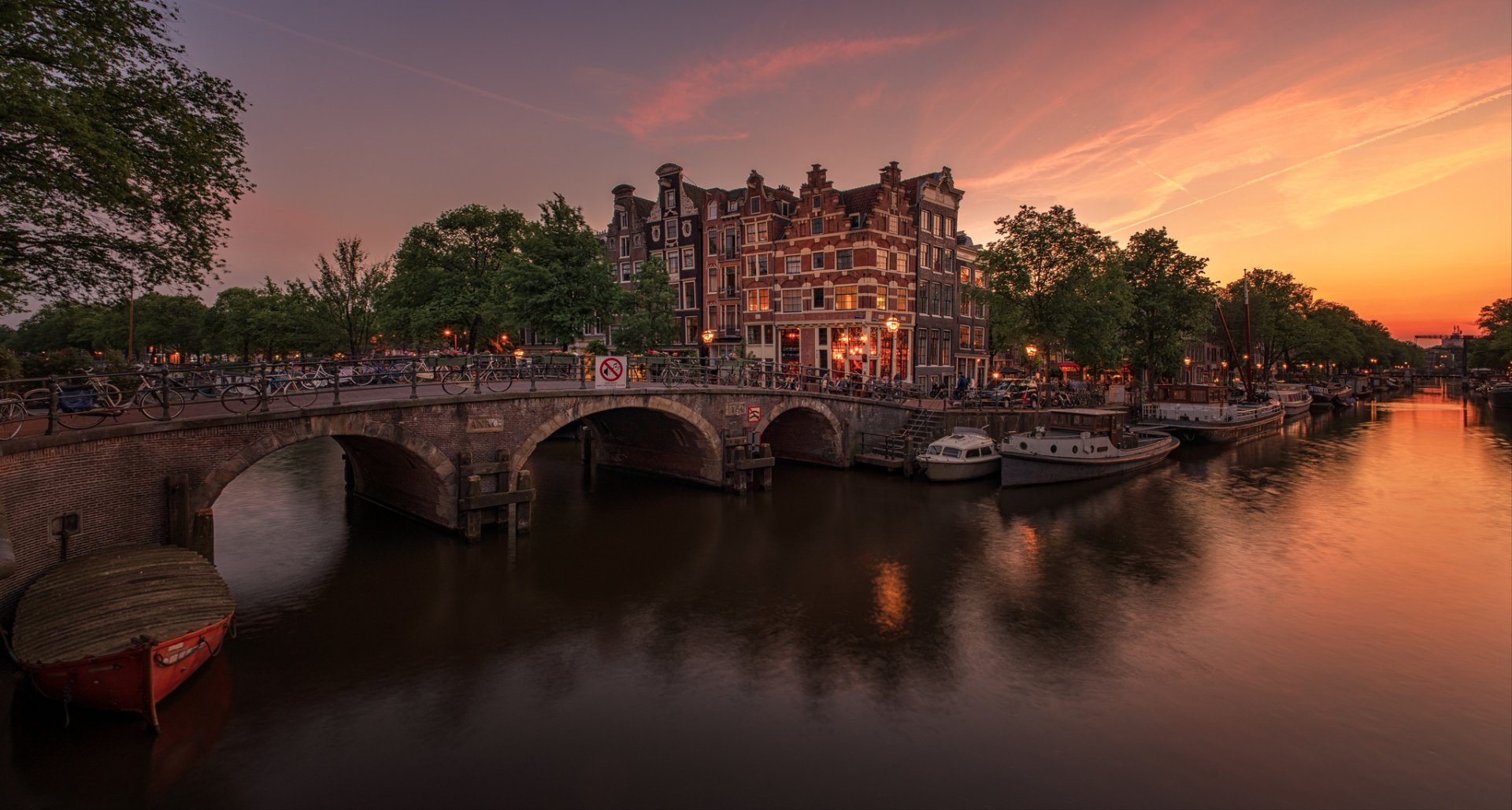 HD desktop wallpaper featuring a man-made canal scene in Amsterdam, Netherlands, with historic buildings and a sunset sky reflecting on calm waters.