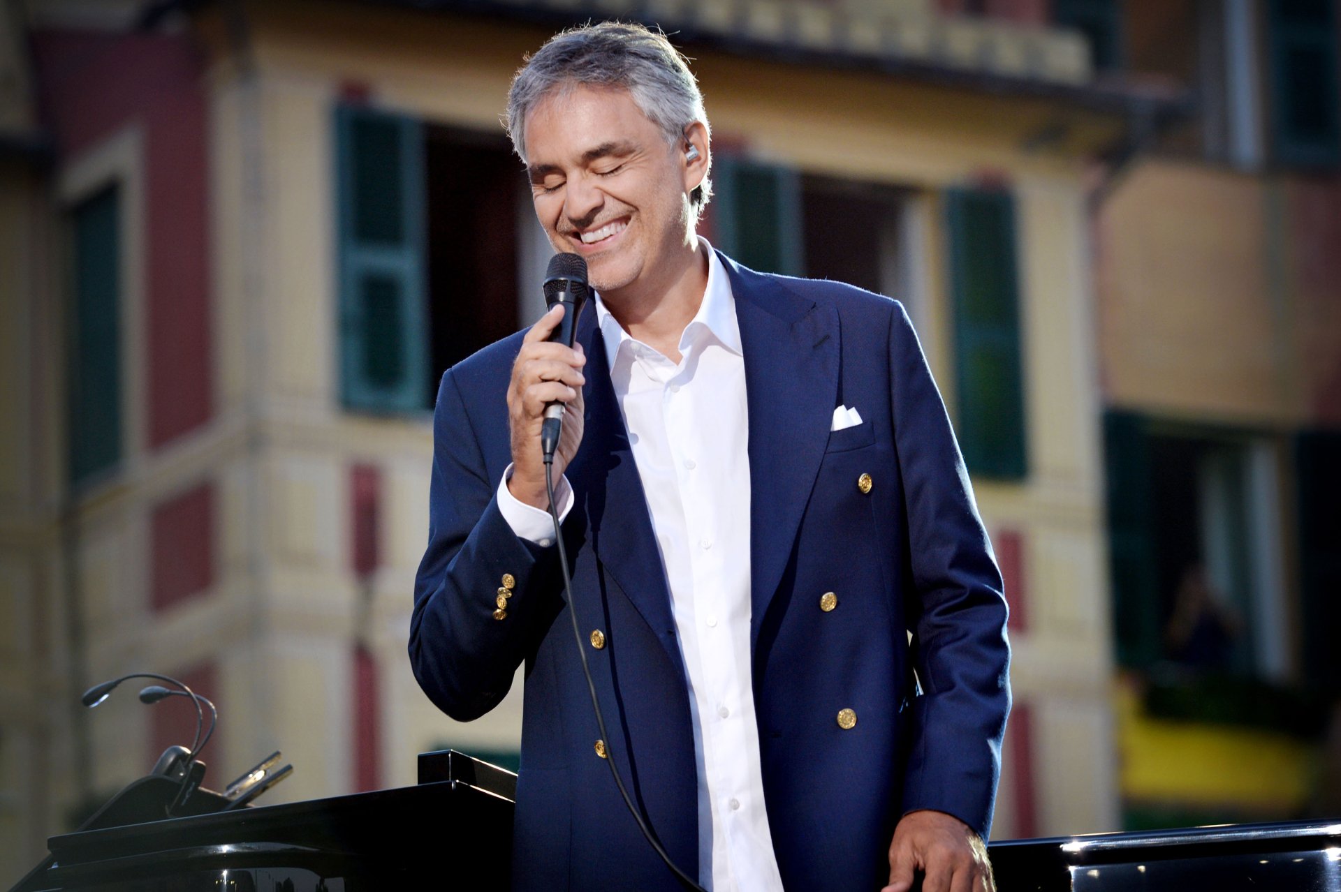 HD wallpaper of a smiling man in a navy blazer holding a microphone, with buildings in the background.