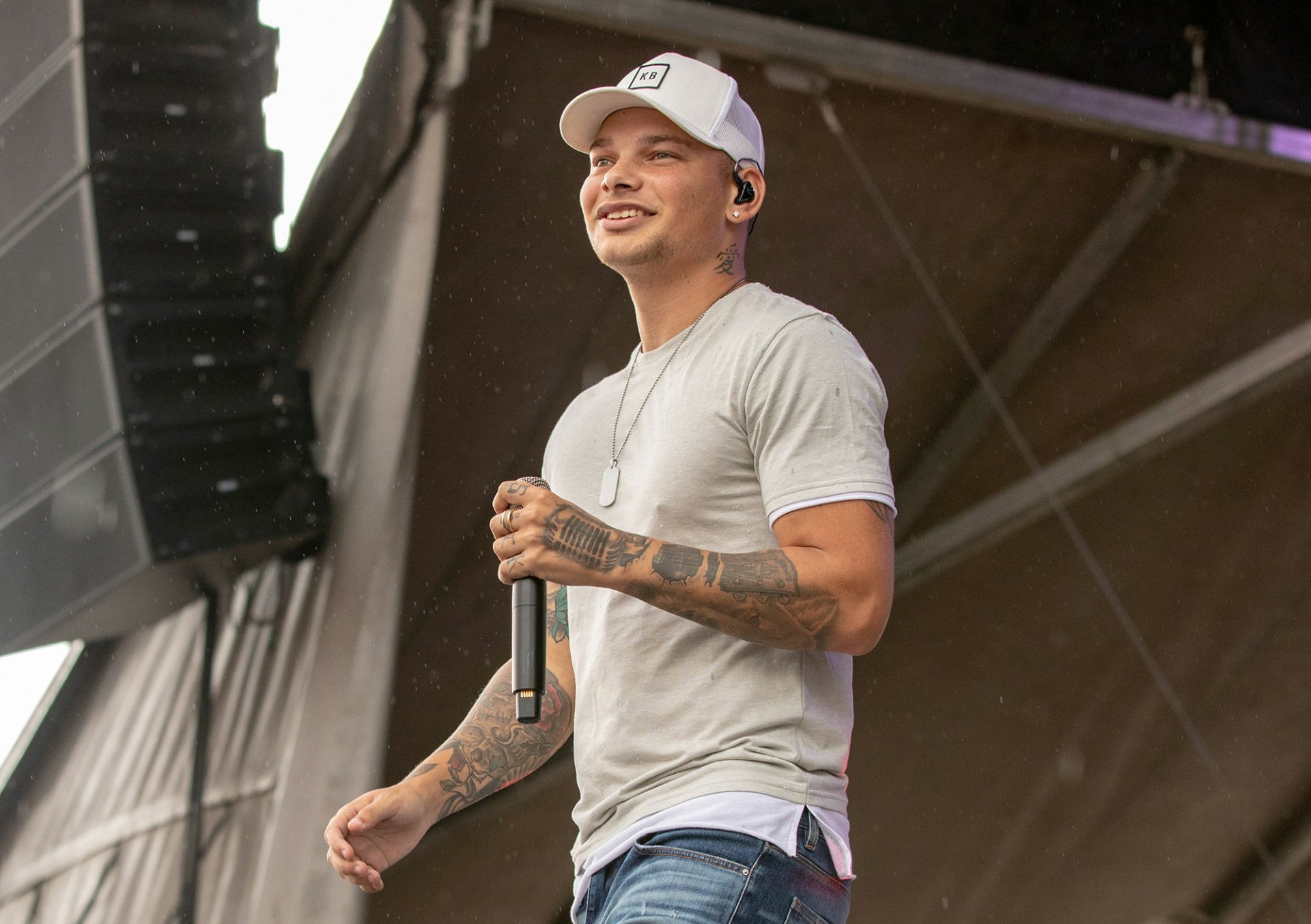 HD wallpaper of a person on stage with a microphone, wearing a white shirt and cap, with tattoos visible on the arms.