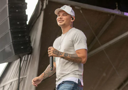 HD wallpaper of a person on stage with a microphone, wearing a white shirt and cap, with tattoos visible on the arms.