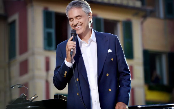 HD wallpaper of a smiling man in a navy blazer holding a microphone, with buildings in the background.