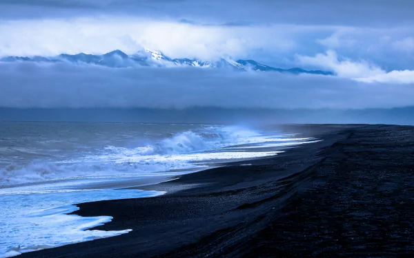 A stunning HD wallpaper of a serene beach with dark sand, gentle waves, and distant snow-capped mountains under a cloudy sky. This nature scene makes for a calming desktop background.