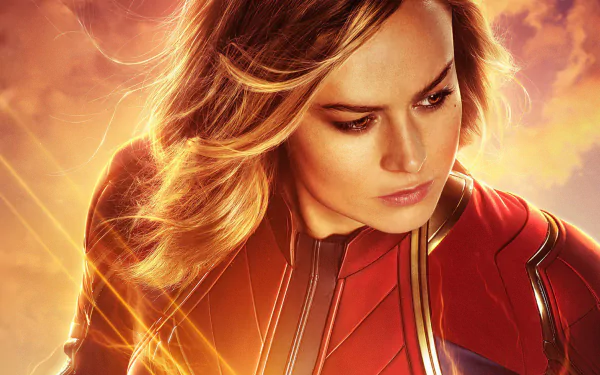 HD wallpaper featuring Brie Larson as Captain Marvel, dressed in a red superhero suit, her blonde hair flowing and brown eyes focused intently. Bright rays of light surround her, creating an intense atmosphere.