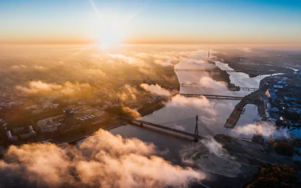 Aerial 5K Ultra HD PC desktop wallpaper of Riga, Latvia, showing a river and cable-stayed bridge through a misty cityscape with the sun on the horizon.