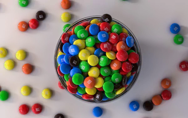 HD PC desktop wallpaper/background of a glass bowl filled with colorful man-made M&M's candy, with more M&M's scattered on a white surface.