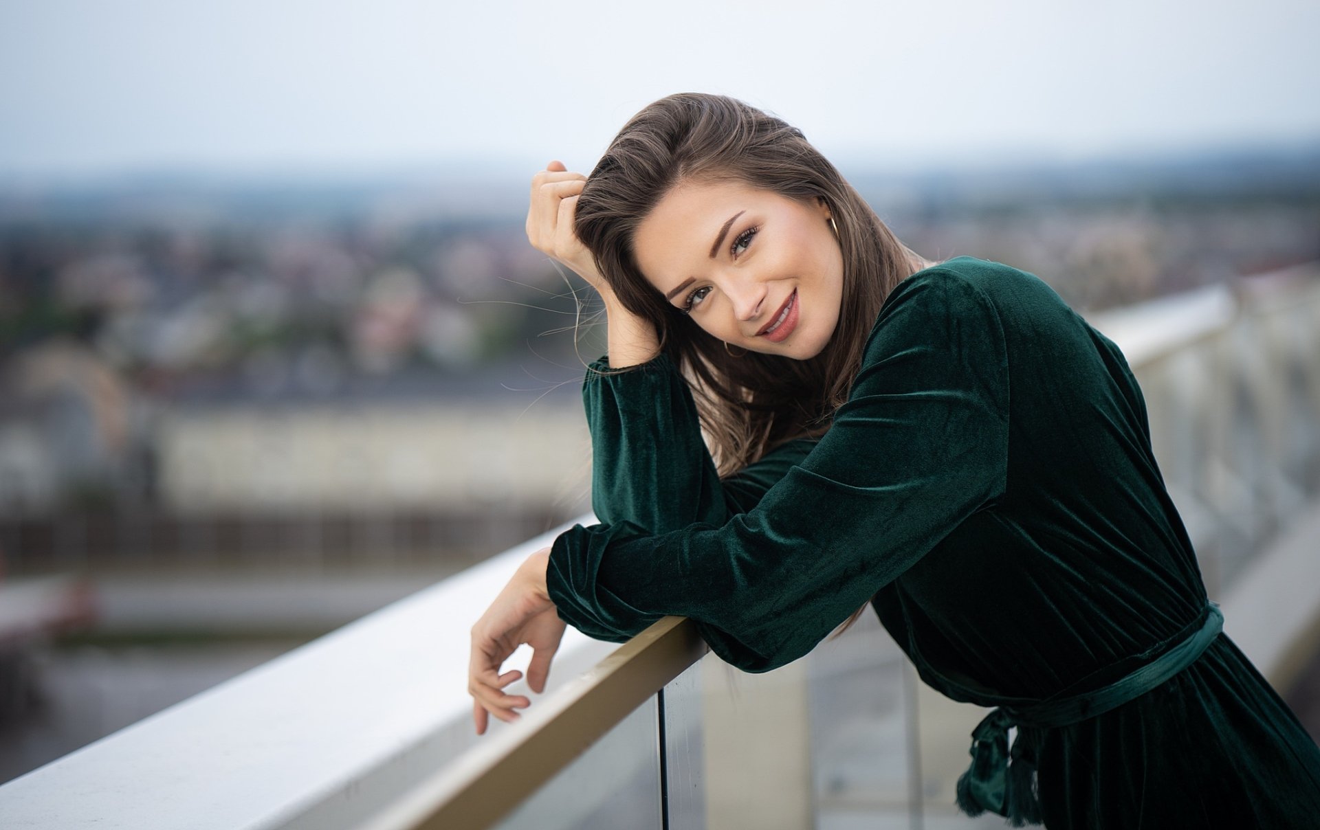 A brunette woman model in a green dress smiles warmly, leaning on a railing with a blurred cityscape in the background, captured with depth of field in this HD desktop wallpaper.
