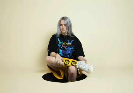 HD desktop wallpaper featuring American singer Billie Eilish seated against a pale yellow background, dressed in a graphic T-shirt and patterned socks.