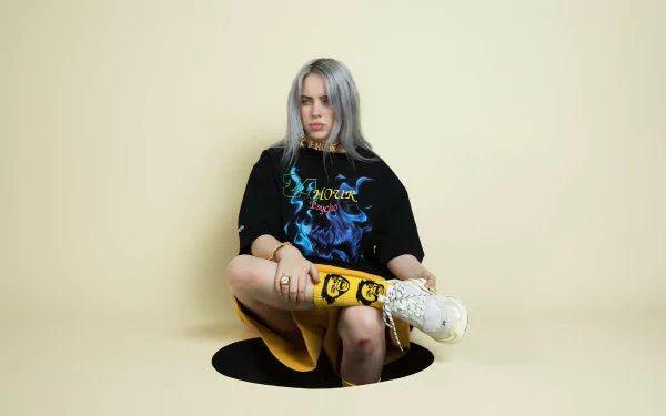 HD desktop wallpaper featuring American singer Billie Eilish seated against a pale yellow background, dressed in a graphic T-shirt and patterned socks.